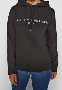 Sweatshirt noir avec capuche, orné d'un logo en blanc et rouge sur le devant. Cordons de serrage avec une finition texturée en or. Coupe décontractée.