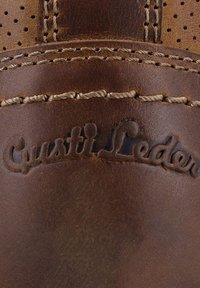 Braune Lederoberfläche mit detaillierter beiger Naht und eingeprägtem, kursivem Text "Gusti Leder" auf dem Leder.