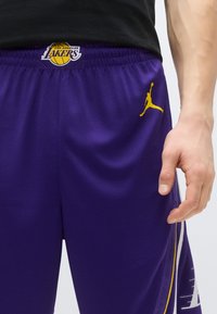 Pantaloni scurți de baschet mov cu logo-ul Los Angeles Lakers și accente galbene, având un material plasă texturat și talie elastică.