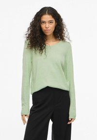 Femme aux longs cheveux bouclés portant un pull à col en V vert clair et un pantalon noir, debout devant un fond blanc uni.