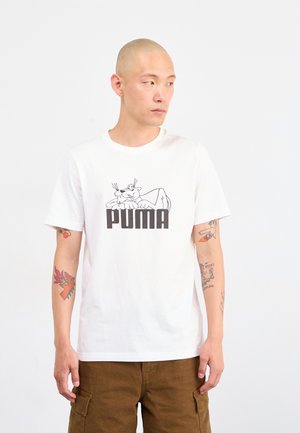 Mann med tatoveringer iført hvit Puma T-skjorte med tegneseriepanterlogo, brune cargo-bukser, stående mot en ensfarget hvit bakgrunn.