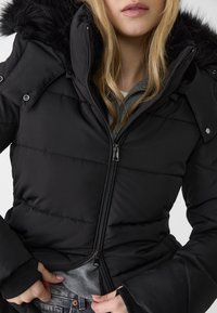 Woman in a black puffer jacket with fur hood, partially zipped, wearing a gray shirt and denim jeans, hands adjusting jacket hem.