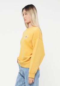 Gul sweatshirt med løs passform, rund hals og lange ermer, med blomsterbroderi foran. Brukes med lyseblå jeans.