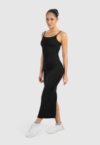 Schwarzes ärmelloses Midikleid in figurnaher Silhouette, mit einem seitlichen Schlitz. Getragen mit weißen Sportschuhen. Glattes, dehnbares Material.