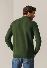 Donkergroene gebreide trui met een textuurafwerking, voorzien van een geribde kraag en manchetten, gecombineerd met lichtblauwe jeans. Zichtbaar achteraanzicht.