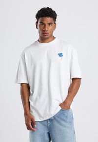 Camiseta de algodón blanca con un corte relajado, que presenta un bordado floral azul en el lado izquierdo del pecho y mangas cortas. Lleva puestas jeans de color azul claro.