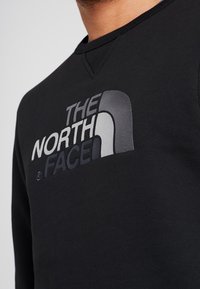 Černá mikina s výrazným šedým a černým potiskem loga "The North Face" na hrudi a kulatým výstřihem. Hladký povrch.