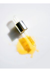 Genomskinlig glasdropperflaska med ett silvermetallock, innehållande en gyllene olja, med en spill på en vit yta.