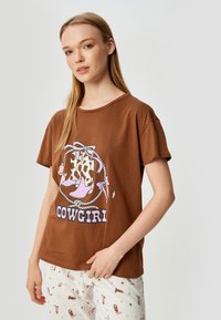 Bruine katoenen T-shirt met korte mouwen, voorzien van een graphic van cowgirl laarzen en touw, en de tekst "COWGIRL" in vette letters.