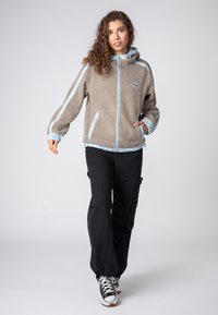 Beige fleecejack met lichtblauwe accenten, een ritssluiting aan de voorkant, zijzakken en een capuchon; gecombineerd met zwarte cargobroek en sneakers.