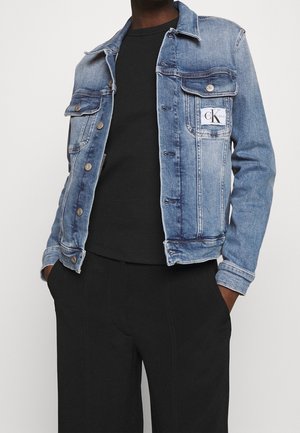 Homme portant une veste en denim bleue avec des boutons et des poches devant, par-dessus une chemise noire et un pantalon noir.