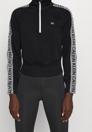 Czarny krótki top z długim rękawem, białym suwakiem na ćwiartkę i logo Calvin Klein na rękawach oraz na piersi, noszony z dopasowanymi czarnymi legginsami.