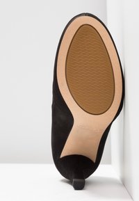 Svart mocka dampumps med spetsig tå och en liten klack. Sulan är beige med strukturerade grepplinjer. Logotyp synlig på sulan.