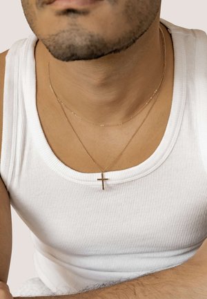 Goldkreuzanhänger mit glatter Oberfläche, geschichtete Ketten, getragen über einem weißen gerippten Tanktop, das einen Nahaufnahme des Ausschnitts zeigt.