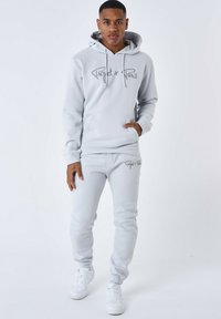 Felpa con cappuccio grigio chiaro e pantaloni jogger abbinati, realizzati in materiale morbido, con tasca frontale e logo testuale. Sneakers bianche.
