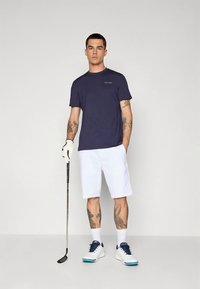 Tamsiai mėlynas trumpomis rankovėmis marškinėlis, balti šortai, balti golfo batai su tamsiai mėlynais akcentais, laikantys golfo lazdelę, stovintys prieš pilką foną.