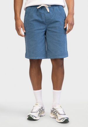 Person iført blå shorts med snøre, hvid t-shirt, hvide strømper og hvide løbesko står mod en lys baggrund.