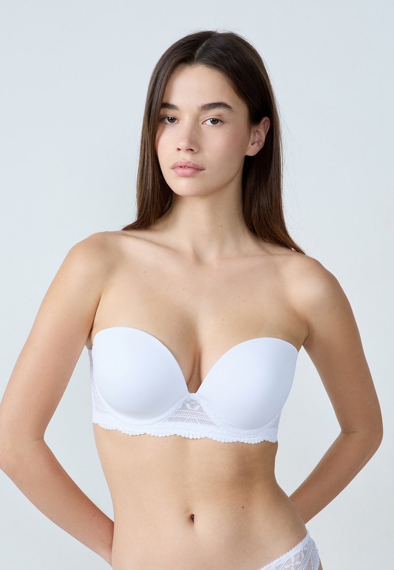 Reggiseno bianco senza bretelle con superficie liscia, rifinitura in pizzo lungo il bordo inferiore e texture morbida. Presenta una coppa a forma di cuore per il sostegno.