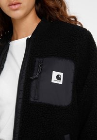 Zwarte fleece jas met een gequilt nylon zak op de linkerkant van de borst met een ritsluiting en een wit logolabel. Soepele voering aan de binnenkant.