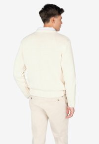 Suéter de punto crema con dobladillo y puños acanalados, llevado sobre pantalones de color claro, mostrando un diseño sencillo y texturizado desde la parte de atrás.