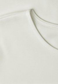 Gros plan d’un tissu blanc montrant des coutures détaillées autour de l’encolure et de la couture d’épaule d’un vêtement doux et lisse.