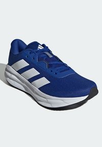 Modrá sportovní bota s síťovaným svrškem, bílým designem se třemi pruhy, polstrovaným límcem a silnou bílou podrážkou. Texturovaná tkanina a šněrovací systém.