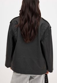 Schwarze Lederjacke mit lässiger Passform, Schulterklappen und sichtbaren Nahtakzenten. Die Oberfläche wirkt glatt und leicht glänzend.