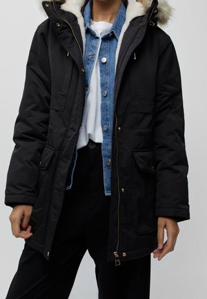 Black parka jacket with faux fur hood, zipper and snap closure, ribbed cuffs, and two front pockets. Worn over a denim shirt and white top.