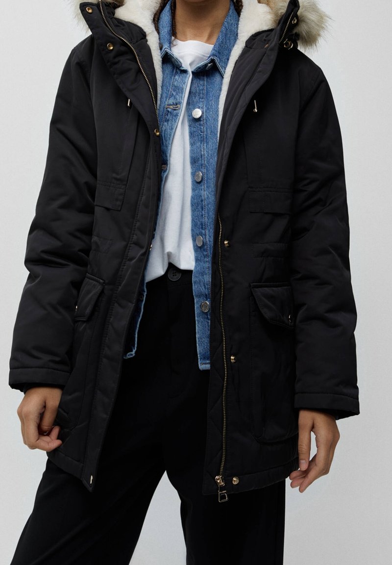Parka nera con cappuccio in pelliccia sintetica, chiusura con zip e bottoni, polsini a costine e due tasche frontali. Indossata sopra una camicia di denim e un top bianco.