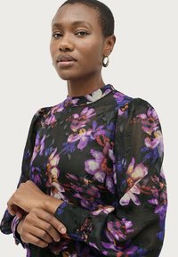 Durchsichtige schwarze Bluse, verziert mit lebhaften lila und pinken Blumenmustern. Hoher Kragen und gepuffte Ärmel mit Knopfmanschetten.