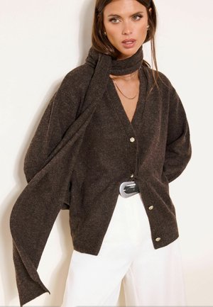 Woman wearing dark brown buttoned cardigan with extended scarf sleeves, white pants, and layered gold necklaces against a plain background.