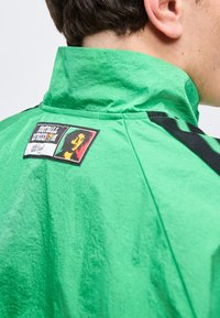 Κοντινό πλάνο ενός ατόμου που φοράει λαμπερό πράσινο μπουφάν με ψηλό γιακά και ένα σήμα που δείχνει ένα πρόσωπο με κόκκινο, κίτρινο και πράσινο φόντο.