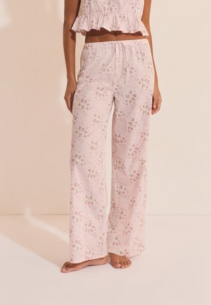 Personne portant un pantalon de pyjama rose clair à petits motifs floraux et un crop top assorti à volants, debout pieds nus sur un sol clair.