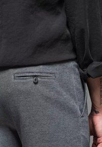Grau gewobene Hose mit flachem Frontdesign und einem Knopfdekor an der Tasche. Der Stoff hat eine strukturierte Optik, die von einem lockeren schwarzen Hemd ergänzt wird.