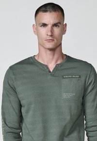 Garā piedurkne zaļa krekls ar textured svītru rakstu, pogātā henley apkakle un krūšu kabata ar drukātām zīmola detaļām.
