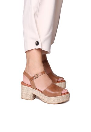 Brown leather platform sandals with a peep toe, featuring an ankle strap and woven jute detailing on the heel, set against light fabric pants.