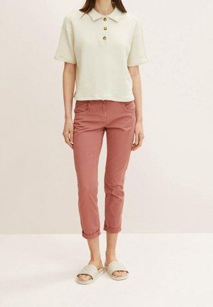 Kobieta w kremowej koszulce polo z krótkim rękawem, zrolowanych różowych spodniach oraz pikowanych beżowych klapkach, stojąca na jednolitym tle.