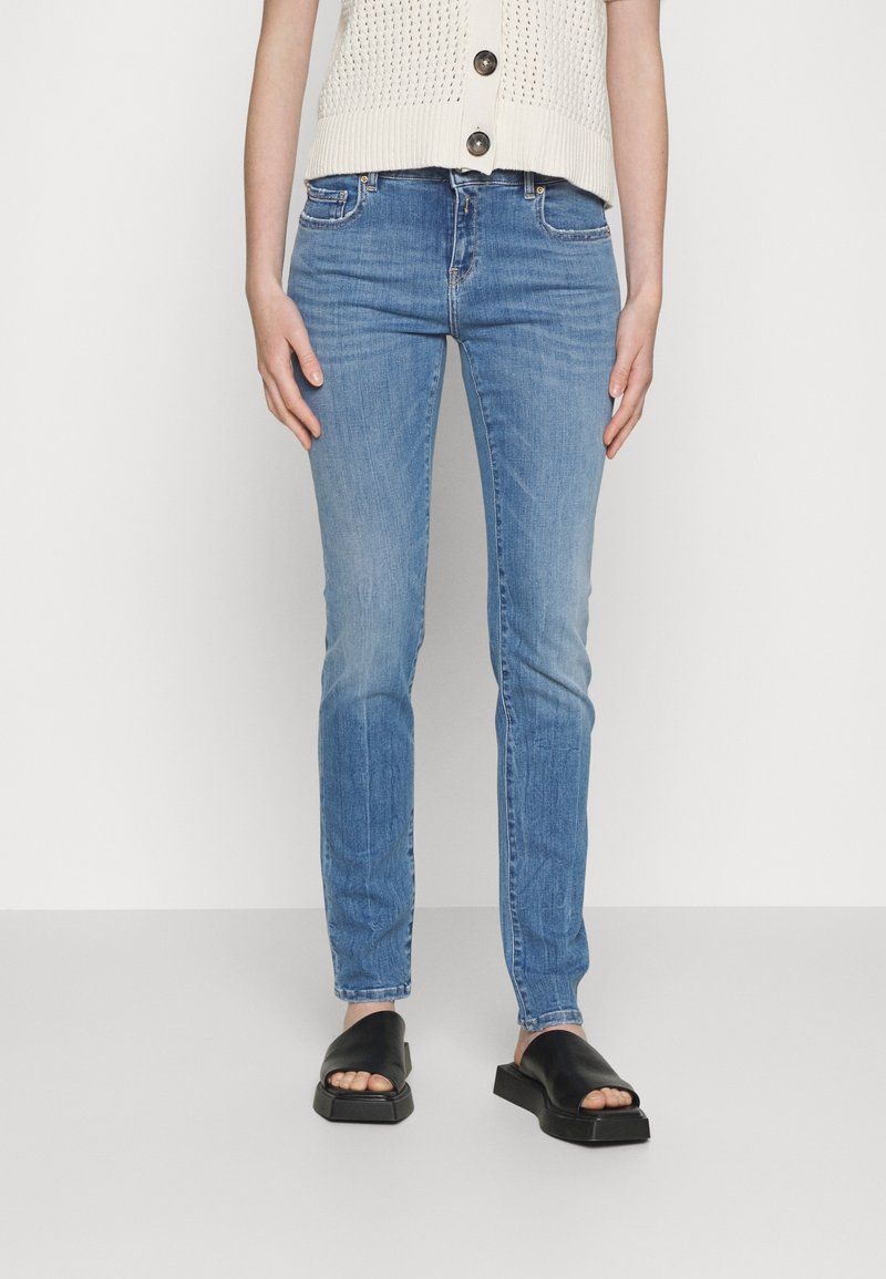 Ljust blå denimjeans med slim fit, lätt blekning på låren, femfickdesign, ihopparade med svarta slidesandaler på en neutral bakgrund.