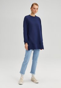 Felpa blu navy con maniche lunghe, colletto rotondo e dettagli a rouches sui lati, abbinata a jeans blu chiaro e sneakers beige.