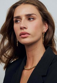 Guldtonade örhängen med cirkulär pärldesign, kompletterade av ett matchande pärlhalsband. Modellen bär en svart blazer. Håret är vågigt och brunt.