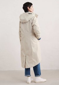 Beige, knie-lange regenjas met een capuchon, twee zijzakken en een rugventilatie, gecombineerd met blauwe jeans en witte sneakers.
