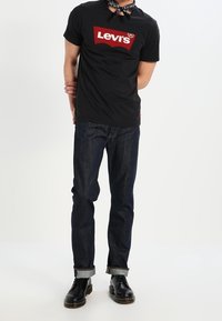 Camiseta negra de manga corta con el logo rojo de "Levi's", combinada con jeans azul oscuro y zapatos de cuero negros con puños enrollados.