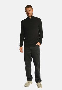 Czarny sweter z krótkim zamkiem, ściągacze w mankietach i u dołu, teksturowana tkanina. Połączony z ciemnymi dżinsami i jasnymi trampkami, stojący na białym tle.