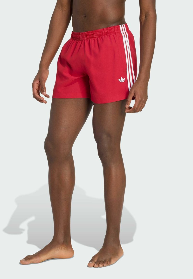 Man wearing red Adidas shorts with white stripes and logo, standing barefoot against a plain light background.