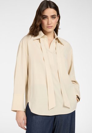 Kvinde med brunt hår iført en løs cremefarvet bluse med lange bindebånd ved halsen og mørkeblå bukser, stående foran en ensfarvet baggrund.