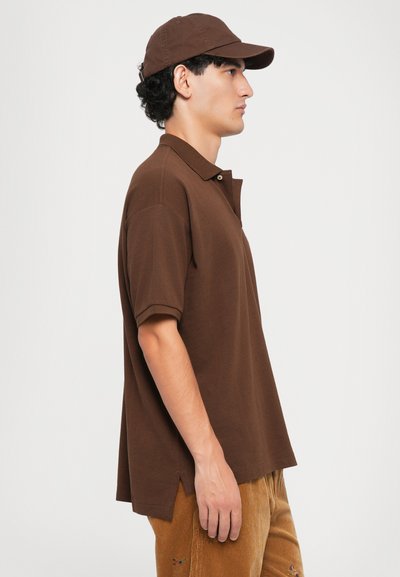 Camisola polo castanha com mangas curtas e colarinho abotoado, usada com calças de veludo cotelê. Um boné castanho a condizer também é usado.