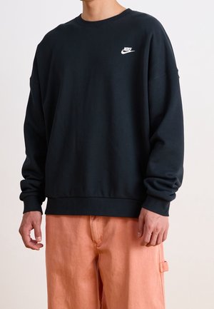 Mann trägt schwarzen Nike-Sweatshirt und lockere pfirsichfarbene Hose, steht vor einfachem hellem Hintergrund.
