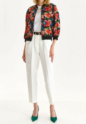 Kobieta ubrana w krótki kwiecisty żakiet, białe spodnie z wysokim stanem, czarny pasek ze złotą sprzączką oraz zielone szpilki, stojąca na białym tle.