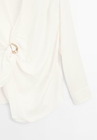 Blusa bianca in seta con un design anteriore annodato e un accento circolare in argento, con maniche lunghe. Texture liscia e vestibilità ampia.