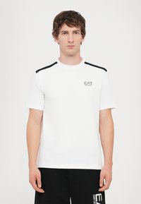 White t-shirt with black shoulder accents and EA7 logo on the chest. Short sleeves, round neckline, made of smooth fabric.